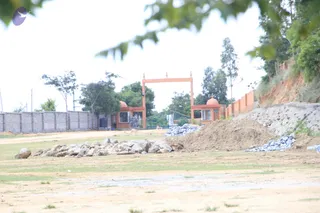 A wide outdoor photo of an unfinished site. In the centre stands an orange-painted gateway with a flat top and small finials, flanked by two small orange domed guard booths with windows. Piles of grey stones and a mound of brown earth lie scattered across dry, patchy grass in the foreground. A grey block boundary wall runs to the left, and a red-earth embankment with a stone retaining wall and green trees rises on the right. No people are visible.