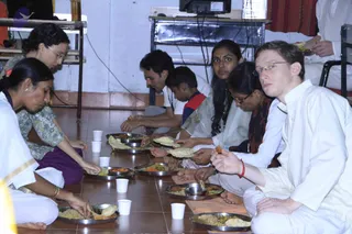 A group of people sit cross-legged on a tiled floor eating a meal from stainless steel plates. A woman in a green floral top and purple skirt on the left ladles food onto a plate, while a woman in white with a gold bangle serves nearby. In the foreground a young man in white kurta with glasses holds food in his right hand, a red thread on his wrist. Others include a woman in a pale sari, a woman in a dark shawl, a man in a white shirt and a boy in a red and blue top. Small white paper cups sit between the plates, and a wall-mounted TV and shelving are behind them.
