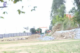 An open dirt and grass ground at a construction site with piles of gravel, rubble and a mound of brown soil. A low grey concrete block wall runs along the left edge, and an orange-painted brick archway frame with rounded domes stands in the background beside trees. A steep embankment of stones and scrubby vegetation rises on the right, with a red brick wall above it. No people are visible in the scene.