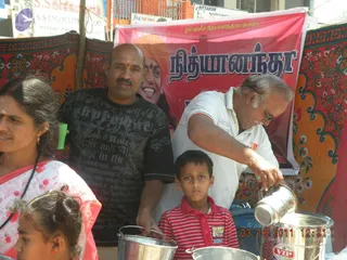 A street-side food distribution scene. A older man in white shirt and glasses pours from a steel vessel into a pot held by a bald man in a black graphic t-shirt. In front stands a boy in a red-and-white striped polo, and at left a woman in a pink-and-white sari with a red forehead mark and a girl are partly visible. Behind them hangs a red banner with a portrait of a smiling bearded figure with a red mark on the forehead, beside the printed name in large letters, and the printed Tamil-language script at top. Steel pots and a large steel container sit in the foreground, with a colourful floral-patterned cloth backdrop.