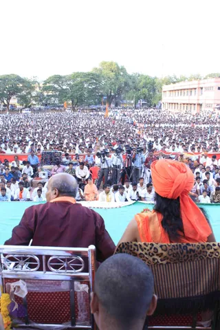 Photograph taken from behind a stage at a large open-air outdoor gathering, with an enormous crowd of men in white shirts filling the ground toward a multi-storey building and trees. In the foreground two seated figures face the crowd: a bald man in a maroon kurta with an orange shawl on the left, and a figure in an orange turban and orange garment with long dark hair on the right, seated in a leopard-print chair. A small shaven-headed child sits in the lower foreground. Video cameras on tripods and a sound mixing setup stand at the stage edge, a row of seated people in orange and white is in front of the crowd, and orange flags are visible in the distance. Marigold garlands are draped near the front chairs.