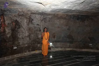 A standing man in a cave temple chamber wears an orange-saffron dhoti and shawl draped over his shoulder, with a long rudraksha mala around his neck. His hair is long and swept back, and he smiles while raising one hand in a gesture of blessing. The rough, rock-walled cave has a dark stone floor, small wall lights, and a low ceiling with cracked rock. A small lamp sits near his feet and a wooden stand is visible beside him. The compact frame shows his posture and clothing in the dim cave light. a person appearance-based label applies only to this standing man with saffron clothing, long swept-back hair and rudraksha mala.
