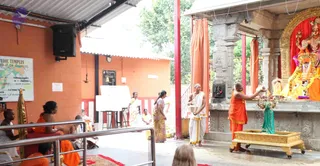 A temple courtyard ritual photographed from the side. A bald priest in white dhoti and shawl stands on the left-centre holding a silver vessel, while a second priest in orange robes at right pours liquid from a small vessel over a figure of a deity on a golden pedestal. Behind them an ornate deity murti with a garland stands in a shrine framed by red curtains and grey stone carvings. Other figures include a woman in a yellow sari and a woman in a pink-and-gold sari standing near a white board, and several seated devotees in orange robes at lower left with a harmonium and a red mat. A wall chart on the left and a small framed sign are visible. The setting is an open-sided temple hall with a corrugated roof and a tree behind.