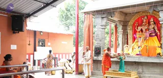 A temple courtyard with orange-washed walls and a red-and-white striped pillar. On the right, a shrine holds large painted deity murtis dressed in red and orange cloth with garlands, a gilded arch behind them. A bald man in an orange robe stands at the shrine offering with a brass vessel, while beside him a priest in white dhoti and shawl holds a small brass pot. Several people in saris and white clothes move around brass pots on the ground, and a woman in a yellow sari with a long braid stands near a whiteboard. Orange curtains hang from the roof and a speaker is mounted on the left wall.