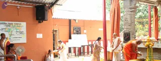 A photograph of an outdoor temple courtyard with orange-painted walls and a tin roof. On the left wall is a map-style sign reading "Nithyananda Vedic Temples" with Kannada text. A shaven-headed man in an orange robe and a standing man in white dhoti stand at right beside a stone shrine with a small deity murti and a brass kalasha-like vessel. A woman in a saree and two men in white dhotis perform ritual actions near a small table with white boards and a fan. A man in white clothing and rudraksha stands near a seated figure at lower left. Orange curtains hang from the roof on the right, and a kumkum-marked stone shrine with carved figures sits behind them.