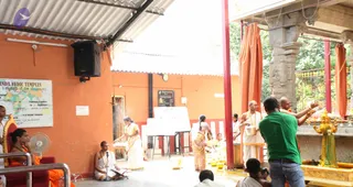 Photograph of a temple ritual in an open orange-walled hall with a corrugated metal roof. A priest in white dhoti with orange shawl and beaded necklaces, seated near a microphone, leads proceedings beside a low red chair. A woman in a white sari with gold border pours from a vessel near a table with a white banner. Another woman in a pink patterned sari stands in the centre. A standing man in a green t-shirt and jeans films or photographs the scene with a camera, while a bald man in white holds up a brass vessel near a golden deity murti on the right. A green-and-red information sign reads Nithyananda Vedic Temples with a world map. Offering pots and a black speaker are visible on the left wall.