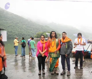 Outdoor photo on a wet stone embankment beside a misty lake with forested hills behind. A long-haired person in an orange and yellow sari-style garment with a green border, red beaded necklace and forehead ornament stands centre, raising a hand. Beside them a woman in a pink top and orange scarf smiles, and a man in grey jacket and orange scarf stands on the right. Another man in white shirt with orange scarf holds a striped umbrella. Several people in the background stand near wooden boats. A woman in orange on the far left wears a red patterned top. Six physical people are visible in total.