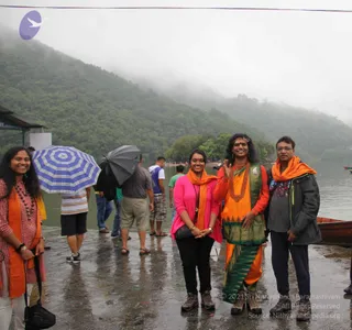 Four people stand on a wet stone lakeside landing under misty, forested hills, with a few boats and other people behind them. Several people hold umbrellas in the background. On the left, a smiling woman wears an orange and red patterned top and beaded necklaces. Next to her, a woman in a pink top and black trousers with an orange scarf stands with hands clasped. In the centre, a figure in a long orange garment with a green and gold bordered wrap has long dark hair, a red bindi and forehead marking, a large red beaded mala, and waves one hand. On the right, a man in glasses wears an orange scarf over a dark grey jacket.